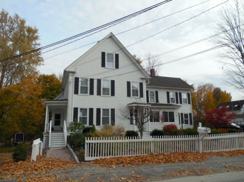 A large white house with black shutters, surrounded by autumn foliage and a white picket fence.