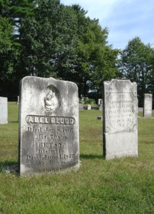 Two weathered gravestones in a grassy cemetery, surrounded by trees, with inscriptions visible on each stone.