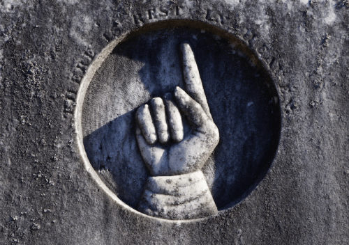 A weathered stone carving of a hand with one finger raised, set in a circular frame.