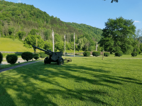 A cannon on a grassy area with trees and flags in the background, set against a hillside under a clear blue sky.