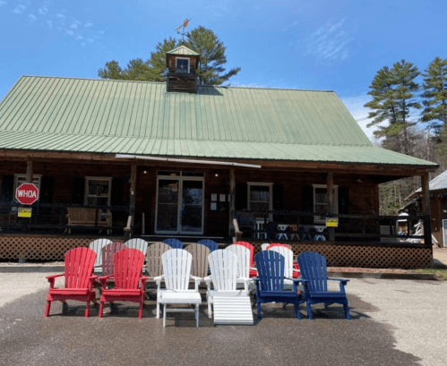 A rustic cabin with a green roof and a porch, featuring colorful Adirondack chairs in red, white, and blue.