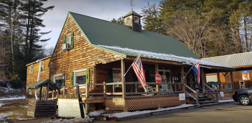 A rustic log cabin with a green metal roof, featuring flags and a porch, surrounded by trees and snow.
