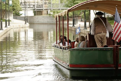 A group of people on a boat in a calm canal, with a man in historical attire steering and an American flag displayed.