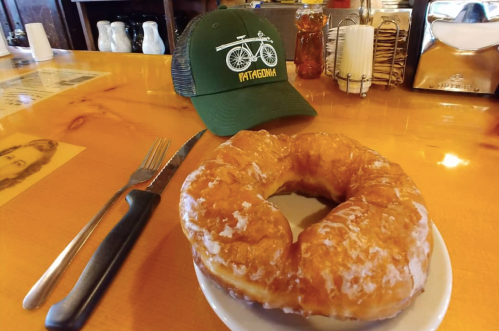 A large glazed donut on a plate next to a fork and knife, with a green Patagonia hat in the background.