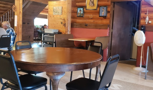 A rustic dining area with a round wooden table, black chairs, and wooden walls, featuring a door and a fan.