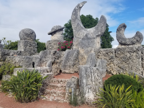 Abstract stone sculptures with circular and crescent shapes, surrounded by greenery and flowers against a blue sky.