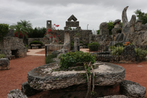 A stone garden with unique rock formations, surrounded by greenery and red flowers under a cloudy sky.