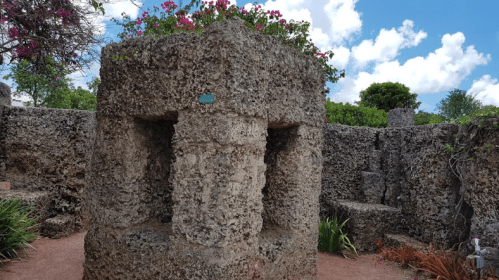 A stone structure surrounded by greenery and flowers, set against a bright blue sky with fluffy clouds.
