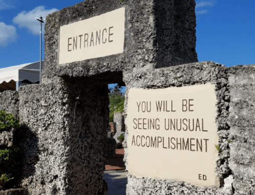 Stone archway with signs reading "ENTRANCE" and "YOU WILL BE SEEING UNUSUAL ACCOMPLISHMENT." Lush greenery in background.