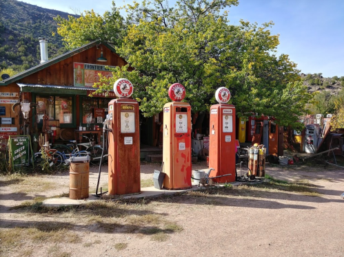 Vintage gas pumps in front of a rustic wooden building, surrounded by trees and old equipment.
