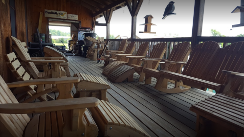 A wooden porch with several Adirondack chairs arranged neatly along the railing, surrounded by a rustic setting.