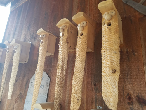 A row of whimsical wooden birdhouses with long, textured bodies and cartoonish faces mounted on a wooden wall.