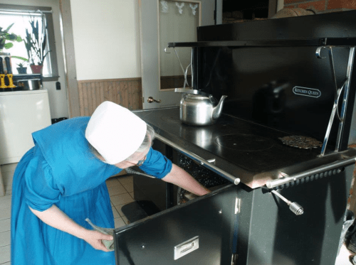 A woman in a blue dress and white bonnet uses a vintage black kitchen stove, reaching into the oven.