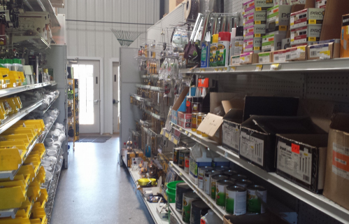 A hardware store aisle filled with shelves of tools, paint cans, and various supplies, with a door at the end.