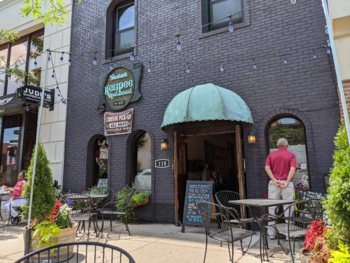 A cozy restaurant entrance with a green awning, outdoor seating, and colorful flowers in pots.