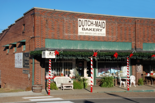 A brick bakery storefront decorated for the holidays, featuring a sign that reads "Dutch-Maid Bakery" and festive garlands.