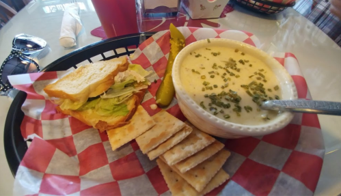 A basket with a sandwich, pickles, crackers, and a bowl of creamy soup on a checkered tablecloth.