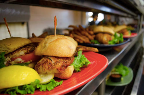 A close-up of a grilled chicken sandwich with lettuce, tomato, and lemon, served on a red plate with fries in the background.