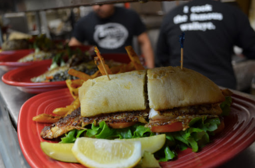 A grilled fish sandwich on lettuce and tomato, served with fries and a lemon wedge on a red plate.