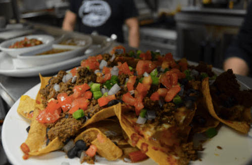 A plate of nachos topped with ground meat, diced tomatoes, onions, and green peppers, served in a kitchen setting.