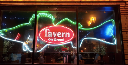 Neon sign of a fish with "Tavern on Grand" illuminated in red, seen through a restaurant window at night.