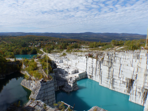 Aerial view of a quarry with white stone cliffs and turquoise water, surrounded by autumn foliage and distant mountains.