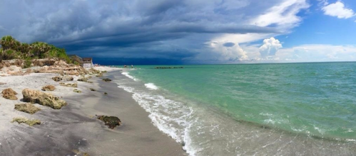 A beach scene with dark storm clouds overhead and clear blue skies in the distance, waves gently lapping at the shore.