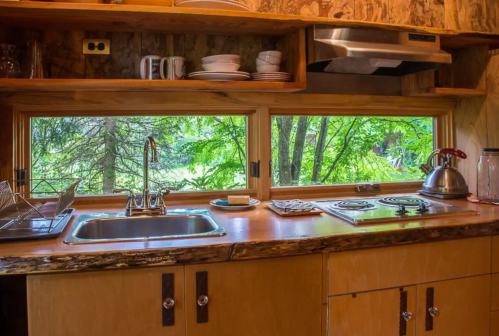 A cozy kitchen with a wooden countertop, sink, stove, and large windows overlooking lush greenery.