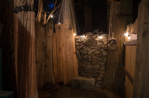 A rustic outdoor shower area with wooden walls, stone backdrop, and string lights, surrounded by trees at night.