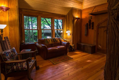 Cozy wooden living room with a leather couch, large windows, and warm lighting, surrounded by nature.