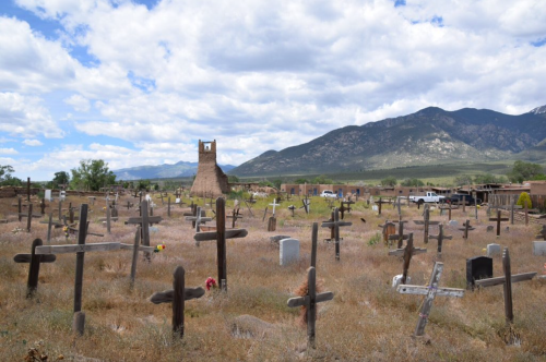 A cemetery with wooden crosses, a stone tower in the background, and mountains under a partly cloudy sky.