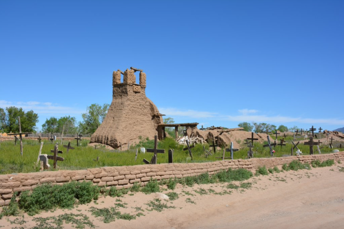 Ruins of a historic adobe structure surrounded by wooden crosses and green grass under a clear blue sky.