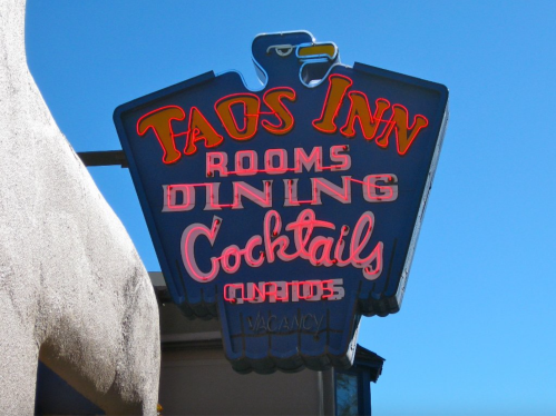 Neon sign for Taos Inn featuring "Rooms," "Dining," "Cocktails," and "Vacancy" against a clear blue sky.