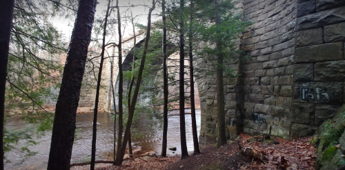 A stone bridge partially obscured by trees, with a river flowing beneath and fallen leaves on the ground.