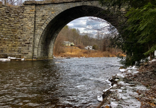 A stone arch bridge spans a river, surrounded by trees and a snowy bank under a cloudy sky.