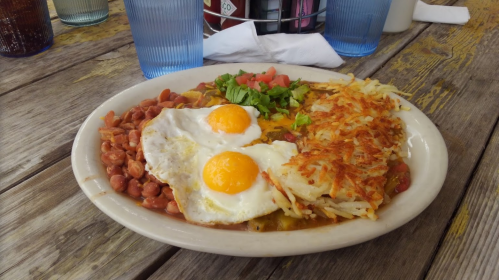 A plate of breakfast featuring two fried eggs, hash browns, beans, and fresh toppings on a rustic wooden table.
