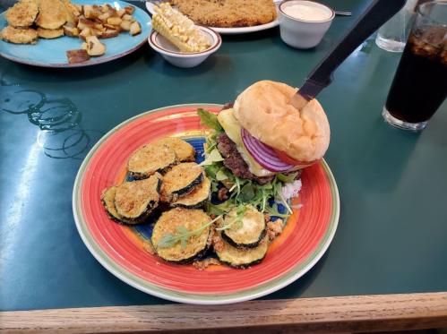 A colorful plate featuring a burger with a knife, surrounded by fried zucchini slices and a side of potatoes.