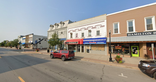 A street view of a small town with shops, trees, and parked cars on a sunny day.