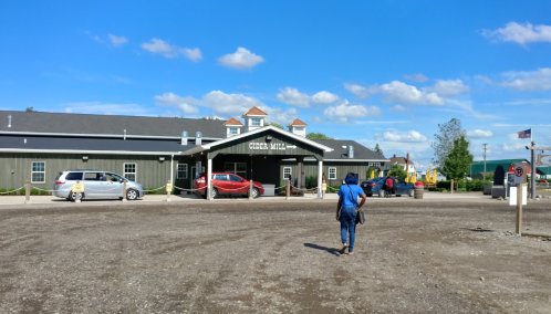 A person walks towards a cider mill building under a blue sky with scattered clouds, surrounded by parked cars.