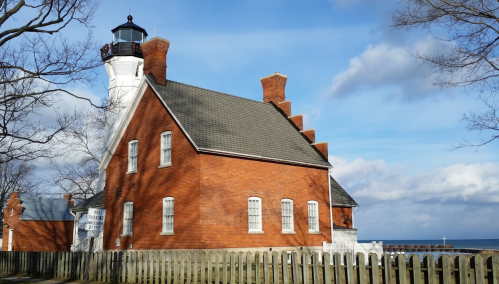 A red brick lighthouse with a white tower, surrounded by trees and a wooden fence, against a blue sky and ocean.