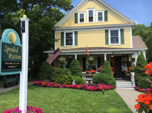 A charming yellow house with green shutters, surrounded by colorful flowers and a sign reading "Spoiled Rotten."