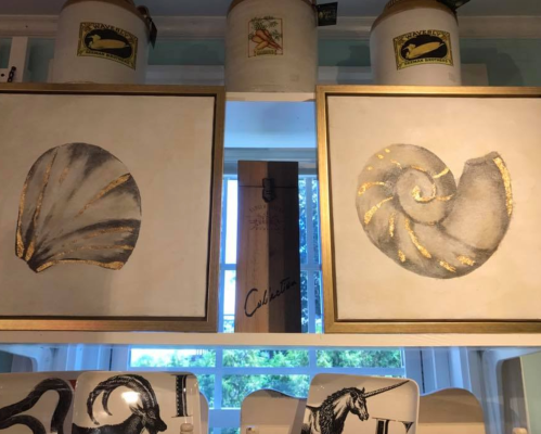 Two framed artworks of seashells with gold accents, displayed on a shelf with decorative containers in the background.