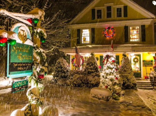 A cozy, snow-covered house decorated for Christmas, with wreaths and a sign for "Spoiled Rotten" in front.