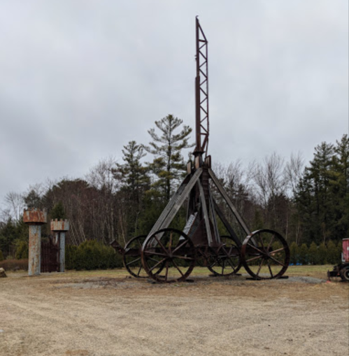 A large, rusted catapult structure stands in an open area, surrounded by trees under a cloudy sky.