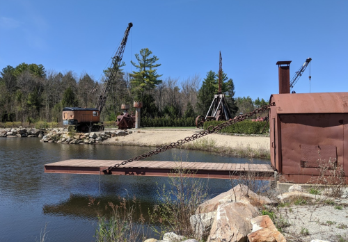 Rusty machinery and cranes by a calm waterway, surrounded by trees and a clear blue sky.