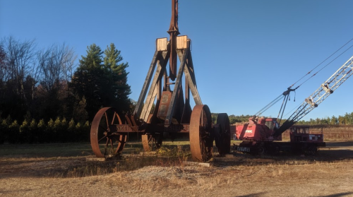 A large, rusted industrial crane stands in a field, surrounded by trees and machinery.