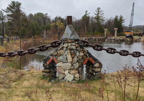 A stone structure with a chain near a calm water body, surrounded by trees and a cloudy sky.