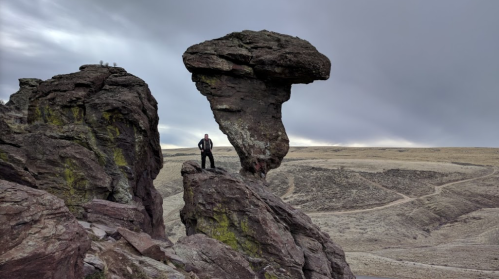 A person stands on a large rock formation resembling a mushroom, with a vast, rocky landscape and cloudy sky in the background.