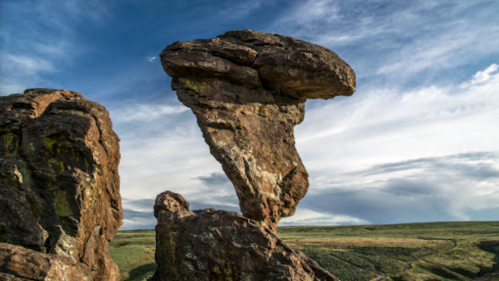 A unique rock formation stands tall against a blue sky with wispy clouds, surrounded by green landscape.