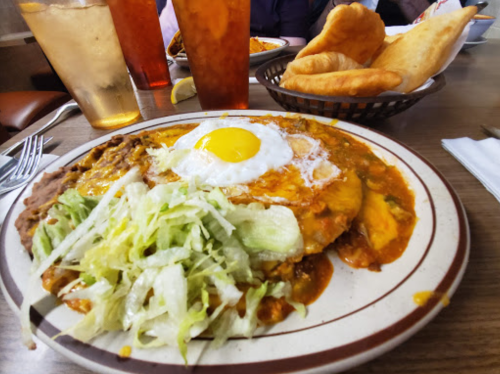 A plate of enchiladas topped with a fried egg, lettuce, and a side of crispy bread, with drinks in the background.